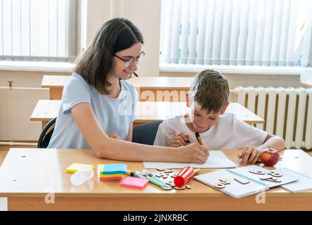 Ein Junge und ein Lehrer machen Hausaufgaben, schreiben Text in ein Notizbuch am Tisch. Eine Mutter hilft ihrem Sohn, in ein Notizbuch zu schreiben. Der Babysitter hilft beim compl Stockfoto