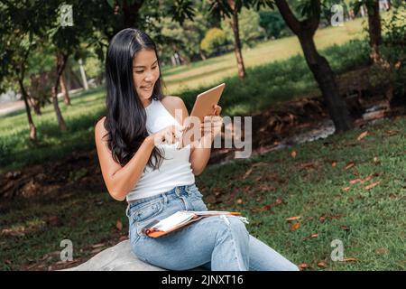 Glückliche Frau, die in einem Park ein Notizbuch schreibt und benutzt Stockfoto