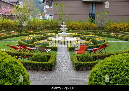 Formeller Garten mit einem Brunnen in der Mitte und gelben Narzissen, die in den Blumenbeeten wachsen. Rote Liegestühle auf dem Gras. Stockfoto