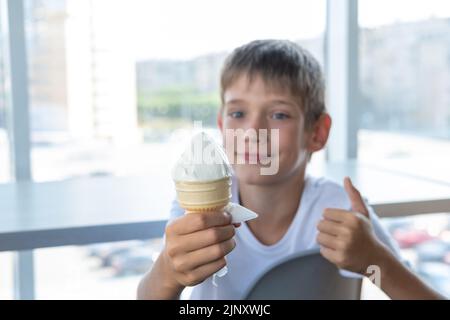 Ein süßer Junge isst weißes Eis in einer Waffelbecher und zeigt eine Daumen-nach-oben-Geste, während er an einem Tisch am Fenster in einem Café sitzt. Unscharfer Hintergrund. A Stockfoto