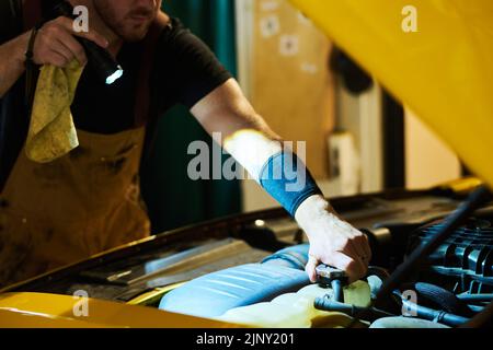 Hand des jungen Mechanikers in der Arbeitskleidung Fixierung Teil des Autos Motor beim Biegen über sie und Regie Taschenlampe auf Motor während der Untersuchung Stockfoto