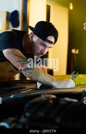 Junge ernsthafte Werkstatt in Arbeitskleidung Biegen nach vorne über offene Haube des Autos und Blick auf den Motor, während sie zu reparieren Stockfoto