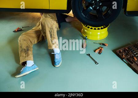 Beine eines jungen Reparaturwerkers oder Technikers in Overalls, die unter einem gelben Elektroauto auf dem Boden einer Werkstatt oder Garae liegen und diese reparieren Stockfoto