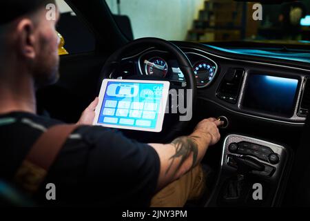 Junger Techniker mit digitalem Tablet, der die Taste auf der Instrumententafel oder auf dem Armaturenbrett des Fahrzeugs drückt, während er vor dem Lenkrad sitzt Stockfoto