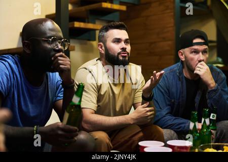 Angespannte interkulturelle Kumpels mit Bier, das am Tisch sitzt und die Übertragung eines Fußballspiels beobachtet, während einer von ihnen etwas sagt Stockfoto