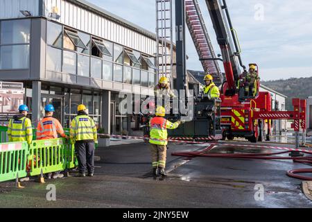 2 die Feuerwehrmannschaft auf einer Hochhubplattform konsultiert einen Kommandanten mit einem Aufzugsbediener auf ihrem Sitz. Eine Bordcrew für Elektrizität schaut nach einer Fabrik Stockfoto