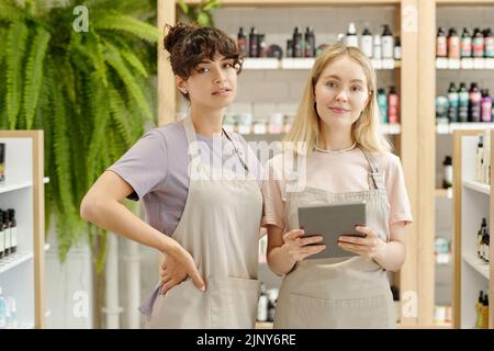 Zwei junge weibliche Angestellte in Schürzen und T-Shirts schauten Sie an, während sie vor der Kamera gegen die Ausstellung im Kosmetikgeschäft standen Stockfoto