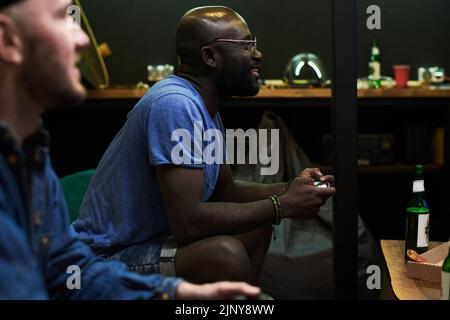 Glücklicher junger afroamerikanischer Mann in blauem T-Shirt, der Videospiel spielt, während er auf der Couch sitzt und Tasten auf dem Gamepad drückt Stockfoto
