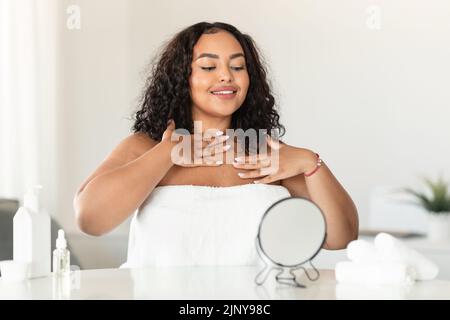 Junge schwarze Plus-Größe Frau berühren Dekolletenzone und Blick in Spiegel, genießen heimische Schönheitsbehandlungen Stockfoto