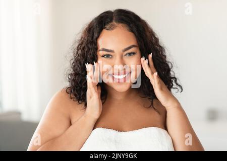 Gesichtsmassage. afroamerikanische Plus-Size-Frau, die ihre glatte, makellose Haut auf den Wangen berührt und die Kamera anlächelt Stockfoto