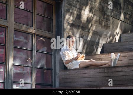Der junge Architekt Freelancer zeichnet ein Bild in einem Skizzenbuch, das auf einer Treppe im Freien sitzt Stockfoto