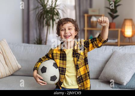 Fußballfan. Glücklicher kleiner Junge, der Fußball trägt und lächelt, um die Kamera zu sehen, die Hand mit Unterstützung hebt, freien Raum Stockfoto
