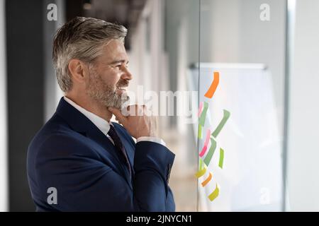 Geschäftsplanung. Reifer Geschäftsmann Liest Notizen Bei Sticky Papers On Glass Wall Stockfoto