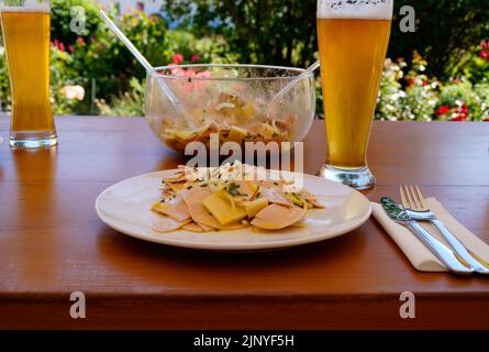 Ein traditionelles, rustikales und herzhaftes deutsches Abendessen in einem Biergarten mit ein paar Bieren, einem Schweizer Wurstsalat mit Käse und Zwiebeln Stockfoto