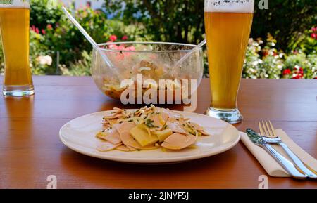 Ein traditionelles, rustikales und herzhaftes deutsches Abendessen in einem Biergarten mit ein paar Bieren, einem Schweizer Wurstsalat mit Käse und Zwiebeln Stockfoto