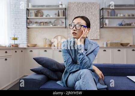 Junge, schöne asiatische Frau, die in einer Brille denkt, die zu Hause im Wohnzimmer auf dem Sofa sitzt, Frau traurig vor Verzweiflung Stockfoto