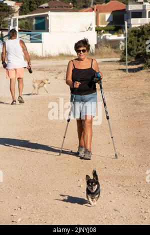 13. Juli 2022: Ältere Frau, die mit ihrem Hund auf einem Feldweg an der Küste mit Spazierstöcken läuft Stockfoto