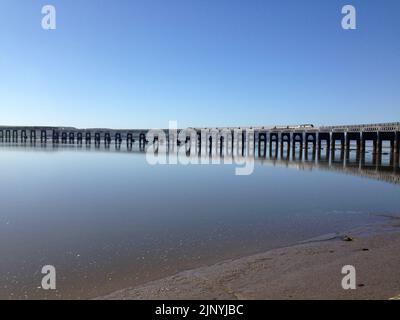 Ein Zug auf der East Coast Mainline, der die Tay Bridge überquert, mit Reflexion im Wasser unter ruhigen Bedingungen unter blauem Himmel in Dundee, Schottland, Großbritannien. Stockfoto