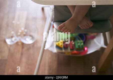 Im Vordergrund sind Babybeine in einem Hochstuhl, im Hintergrund ist eine zerbrochene transparente Platte. Das Konzept der Küchenunfälle ist für Kleinkinder gefährlich. Hochwertige Fotos Stockfoto