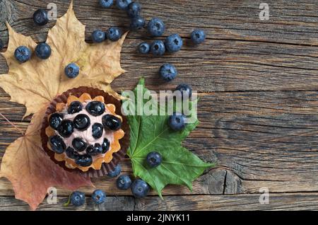 Käsekuchen mit frischen Blaubeeren und herbstlichen Ahornblättern auf altem Holzhintergrund, Draufsicht mit Platz für Text Stockfoto