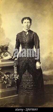 Ende 19. Century Photo Studio Porträt einer formellen ernsthaften Frau, die in stilvoller, zeitlicher Kleidung mit Hintergrund um 1870 - 1880s steht Stockfoto