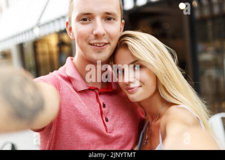 Lächelnd schönes Mädchen und ihr Freund in lässigen Sommerkleidung. Glückliche Familie, die Selfie-Selbstporträt von sich selbst auf der Smartphone-Kamera. Spaß auf der Straße Hintergrund. Stockfoto
