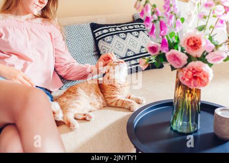 Spielen mit Katze zu Hause. Junge Frau, die sich auf der Couch im Wohnzimmer entspannt und Kätzchen mit Blumen in der Vase streichelt. Sich wohlfühlen und wohlfühlen Stockfoto