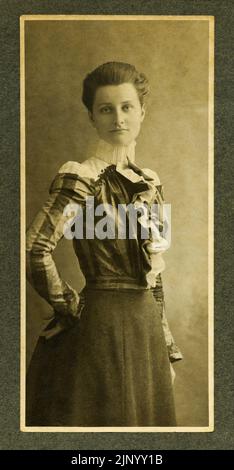 Vintage-Fotografie, aufgenommen Ende des 19.. Jahrhunderts, mit einer schönen jungen Frau, die direkt in die Kamera blickt und stilvolle historische Kleidung aus der viktorianischen Zeit um 1890 trägt Stockfoto