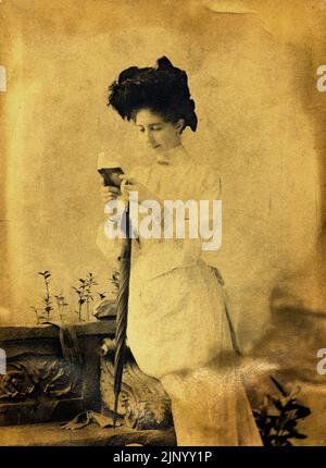 Vintage-Fotografie, aufgenommen im Porträtstudio Ende des 19.. Jahrhunderts, bei der junge Frau in historischer Kleidung saß und ein Buch las und einen Regenschirm hielt, während sie um 1889 für die Kamera posierte - Budapest Stockfoto