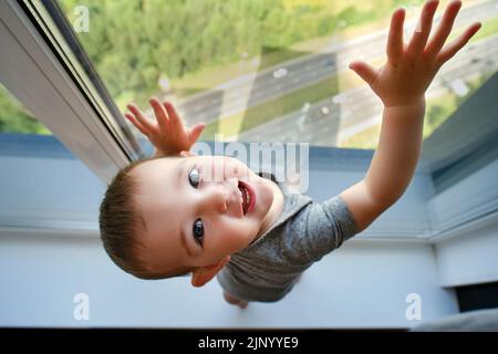 Kleinkind Baby steht am Fenster auf der Fensterbank. Kind am Fenster, das die Hände am Glas hält. Kind im Alter von einem Jahr Stockfoto