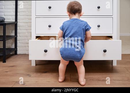 Kleinkind Baby öffnet eine Kommode. Der Junge greift in eine offene Schublade eines weißen Schranks. Kind im Alter von einem Jahr Stockfoto