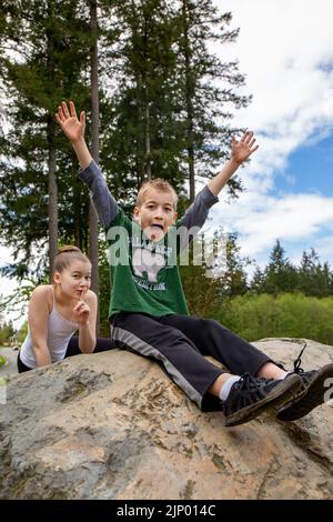 Issaquah, Washington, USA. Die 11-jährige Schwester schleicht sich hinter ihrem 9-jährigen Bruder, der dumm und brüllend auf einem Felsblock in einem Park sitzt. Stockfoto