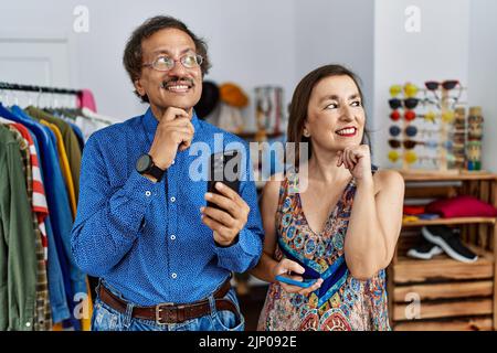 Interracial Paar im mittleren Alter im Einzelhandel mit Smartphone mit Hand auf Kinn denken über Frage, nachdenklichen Ausdruck. Lächelnd und nachdenklich fa Stockfoto