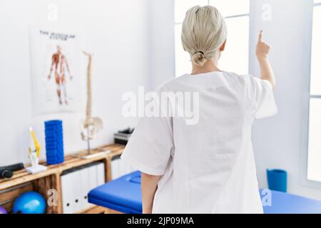 Junge kaukasische Frau, die in der Schmerzklinik arbeitet, posiert rückwärts und zeigt mit der Fingerhand nach vorne Stockfoto
