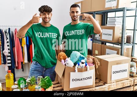 Junge schwule Paar trägt Freiwilligen T-Shirt bei Spenden stehen mit Hand Finger auf Gesicht und Nase, lächelnd fröhlich. Beauty-Konzept Stockfoto