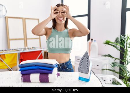 Junge hispanische Frau bügelt Kleidung in der Waschküche tut ok Geste wie Fernglas aufragende Zunge aus, Augen durch die Finger schauen. Crazy Express Stockfoto