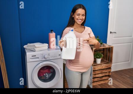 Junge, schwanger Frau, die Wäsche hält, hält die Waschmittelflasche lächelnd und zeigt glücklich mit Hand und Finger Stockfoto