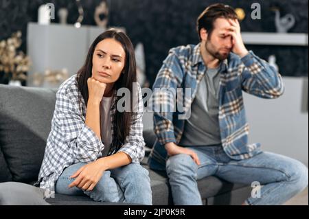 Ein Streit in der Beziehung eines kaukasischen Paares, die Ehepartner zanken über Meinungsverschiedenheiten, sie sitzen auf einem Sofa im Wohnzimmer, sie werden gegenseitig beleidigt, sie schauen in verschiedene Richtungen Stockfoto