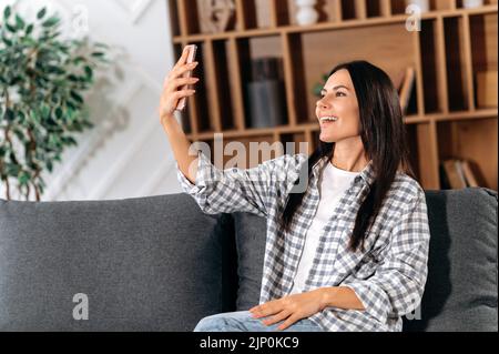 Positive Mädchen sitzen auf dem Sofa im Wohnzimmer, nutzt Smartphone, um per Videokonferenz mit Freunden zu kommunizieren, Gruß, Lächeln. Glückliche kaukasische junge Frau in legerer Kleidung, nimmt ein Selfie am Telefon Stockfoto