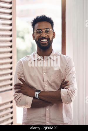 Glücklicher, sorgloser und positiver junger Geschäftsmann, der bei der Arbeit in seinem Büro mit gekreuzten Armen steht. Porträt eines lächelnden männlichen Mitarbeiters mit Brille Stockfoto