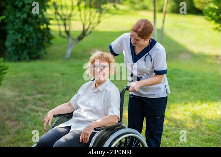 Kaukasische Ärztin geht mit einer älteren Patientin im Rollstuhl in den Park. Krankenschwester begleitet eine alte Frau auf einem Spaziergang im Freien. Stockfoto