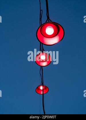 Drei Lampen hängen an Kabeln vor einem blauen Abendhimmel. Aus den Glühbirnen leuchtet rotes Licht. Eine abstrakte Szene mit roten Straßenlichtern. Stockfoto