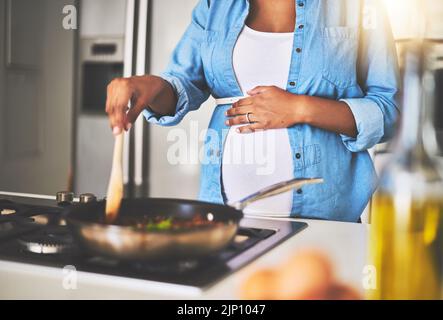 Kochen bis zu ihrem letzten Trimester. Eine Schwangere bereitet zu Hause eine Mahlzeit auf dem Herd zu. Stockfoto