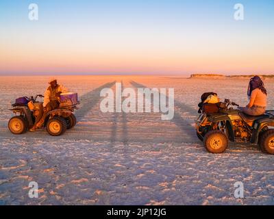 Der Sonnenuntergang erhellt die Wüstenlandschaft und zwei Menschen auf Quad-Bikes während eines Abenteuers in der Makgadikgadi Desert Pan in Botswana. Stockfoto