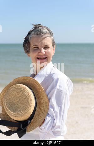 Fröhliche lächelnde Frau in einem weißen Herrenhemd, die einen Bootshut mit einem schwarzen Band in den Händen hält, vor dem Hintergrund des Meeres und des blauen Himmels. Stockfoto