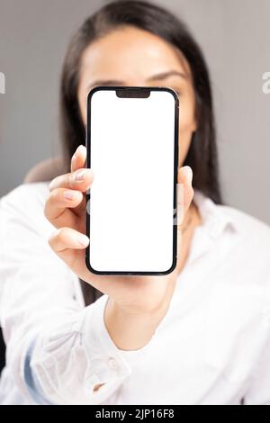 Die junge Frau hält das Smartphone mit dem Smartphone vor ihrem Gesicht. Empfehlungskonzept. Neues Mobiltelefon mit weißem leeren Bildschirm für Mockup. Stockfoto