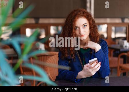 Portrait glücklich lächelnd hübsches rothaariges Mädchen, das im Straßencafé oder Co-Working-Space sitzt, Mobiltelefon benutzt, SMS schreibt oder Live-Übertragungen anschaut, Online-Webinar, Selbstschulung mit einer nützlichen Anwendung Stockfoto