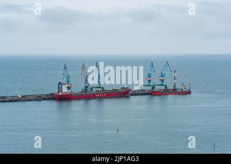 Korsakov, Russland - 04. August 2022: Frachtliegeplatz mit Hafenkranen und festgetäuten Schiffen vor dem Hintergrund des offenen Meeres Stockfoto