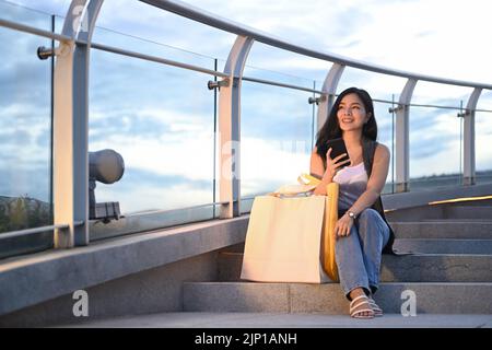 Trendige junge asiatische Frau mit Einkaufstaschen auf Treppen mit Hintergrund der Stadtlandschaft bei Sonnenuntergang sitzen Stockfoto
