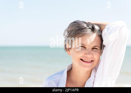 Glücklich lächelnde grauhaarige Frau in einem weißen Männerhemd, die sich mit der Hand gegen das Meer und den blauen Himmel vor der Sonne schützt. Stockfoto